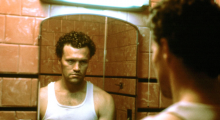 A man in a white undershirt stares into a grimy mirror.